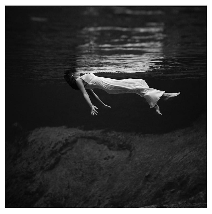 Woman floating above a large rock in water wearing a long white dress.