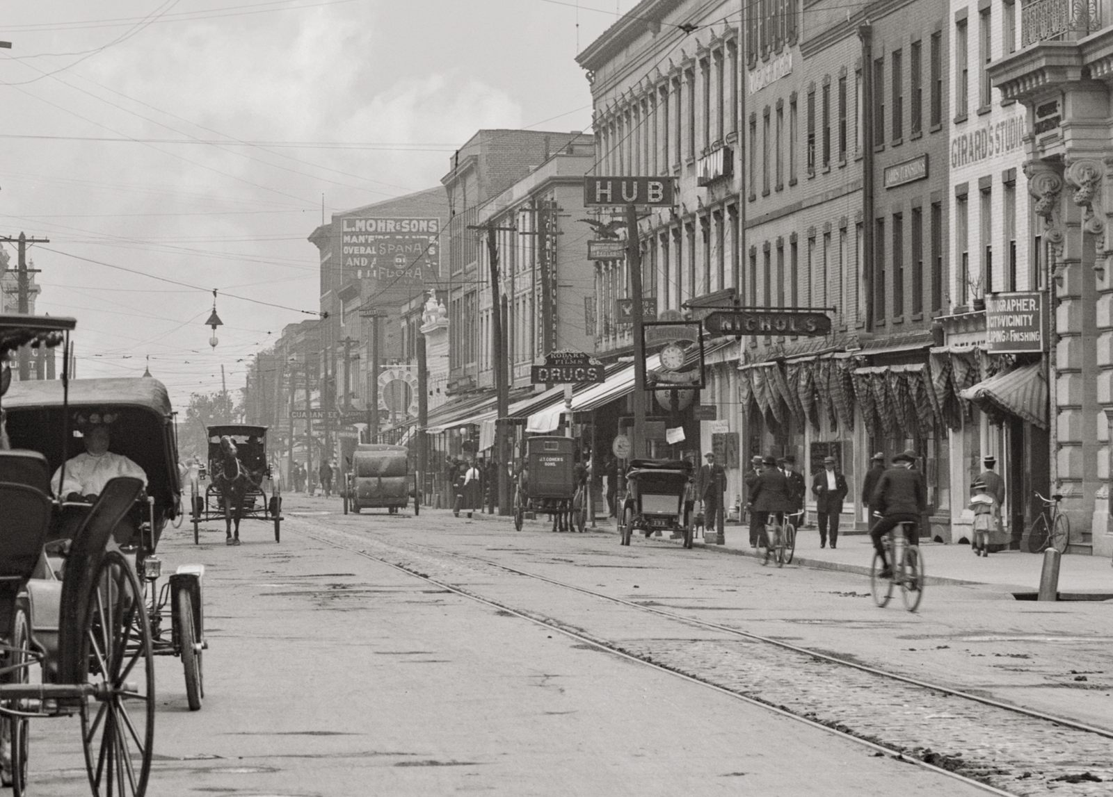 A closeup of the street with people riding bicycles and horse drawn buggies.
